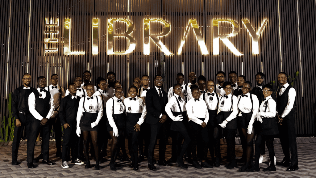 A group photo of The Library Lagos service team — bartenders, waiters, and waitresses — all dressed in coordinated black and white uniforms during customer service week, standing confidently in front of The Library’s illuminated exterior signage.